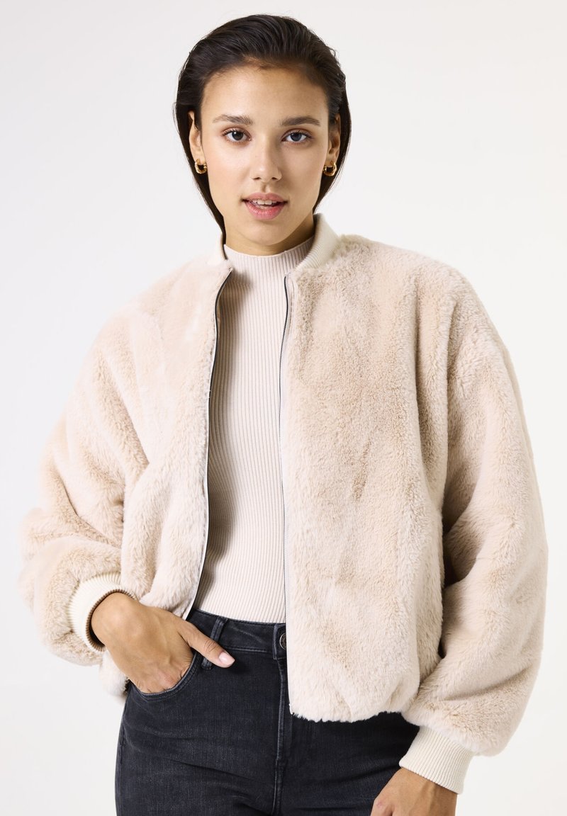 La veste bomber en fausse fourrure beige présente une fermeture éclair, des bords-côtes et une coupe décontractée. Portée par-dessus un haut léger en côtes et un jean sombre.