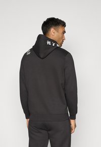 Sweatshirt à capuche noir en tissu texturé. Présente un logo sur la capuche et des poignets côtelés. Design simple avec une coupe décontractée.