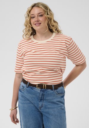 Frau mit lockigem blondem Haar, die ein cremefarbenes und orange gestreiftes Kurzarmshirt, blaue Jeans, schwarzen Gürtel und ein goldenes Armband trägt, steht mit der Hand in der Hüfte.