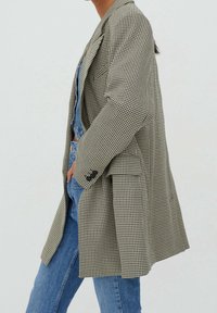 Blazer à carreaux noir et blanc, dot é d'une coupe décontractée, de manches longues et de poches latérales. Porté par-dessus une salopette en denim et un jean bleu.