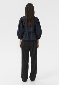 Marineblusiertes Peplum-Top mit Puffärmeln, kombiniert mit schwarzen weit geschnittenen Hosen. Glatte Textur; Knöpfe im Rücken.
