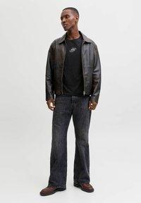 Czarna skórzana kurtka, czarny t-shirt z logo, ciemne dzwony z denimu, brązowe buty. Model stoi z rękami luźno opuszczonymi na neutralnym tle.