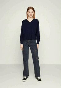 Navy V-hals trui met ribbeltextuur, gecombineerd met donkere cargobroeken met zijzakken en zwarte sneakers met witte zolen.