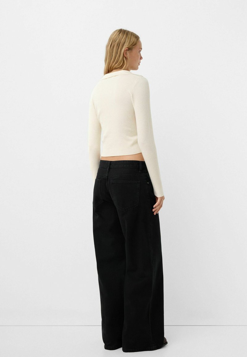 Top a maniche lunghe a costine color crema abbinato a jeans neri a gamba larga a vita alta, che mettono in risalto una silhouette aderente e un tessuto strutturato.