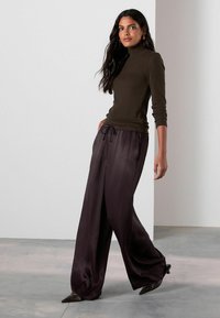 Ciemnozielony dopasowany golf zestawiony z luźnymi, błyszczącymi, szerokimi spodniami w ciemnym odcieniu fioletu. Modelka ma na sobie czarne spiczaste buty.