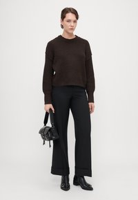 Brown knitted sweater with a crew neck, paired with black wide-leg trousers. The model holds a small black handbag. Textured fabric details visible.