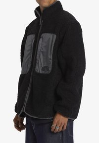 Svart fleecejacka med grå nylonfrontpaneler, hög krage, blixtlåsstängning och textilstruktur. Med två framfickor och ribbade ärmslut.