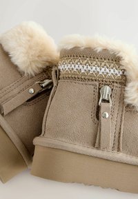 Beige Knöchelstiefel aus Wildleder mit einem Kunstpelzbesatz, einer dekorativen geflochtenen Borte und metallischen Hardware-Details. Strukturierte Sohle für besseren Halt.