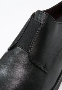 Chaussure en cuir noir avec panneau élastique sur les côtés, bout arrondi, texture lisse et détails de couture minimalistes. Convient à un usage décontracté ou formel.
