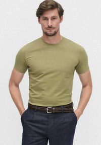 Olijfgroen T-shirt met korte mouwen, ronde hals, aansluitend ontwerp, gecombineerd met donkere broeken en een gevlochten bruine leren riem. Glad textuur.