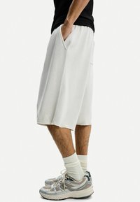 Witte knielange shorts met een elastische tailleband, zijzakken en een rafelige zoom, gedragen met grijze en beige sportschoenen en witte sokken.