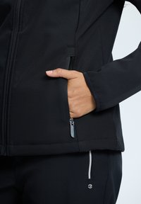 Giacca softshell nera con tasca con zip e texture liscia. Il design presenta una forma aderente con cuciture dettagliate.