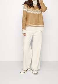 Brun stickad tröja med ett vitt diamantmönster, kombinerad med vida vita byxor och ljusa skor. Enkel, strukturerad design.