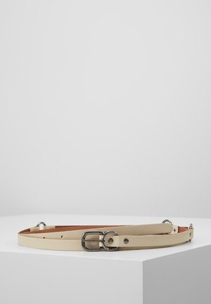 Beige leren riem met zilverkleurige gesp en metalen ringen, plat gelegd op een witte, schuine ondergrond tegen een effen achtergrond.