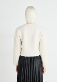 Krämfärgad, fluffig kort jumper med långa ärmar, slät textur och synliga sömdetaljer, i kombination med en svart plisserad kjol.