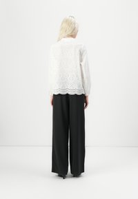 Blusa bianca di pizzo con orlo scallop, caratterizzata da dettagli perforati. Abbinata a pantaloni neri a vita alta e a gamba larga.