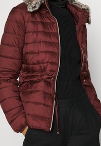 Personne portant une veste matelassée bordeaux avec capuche en fourrure, ouverte sur un pull en maille noir et un pantalon noir, les mains dans les poches.