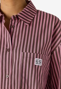 Brun og rosa stripete skjorte med knappeslukning på kragen, liten lomme og en logo-patch. Jevn tekstur og rette vertikale linjer.