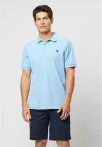 Šviesiai mėlynas polo marškinėliai su trumpomis rankovėmis ir apykakle, dekoruoti juodu logotipu krūtinėje. Suderinti su tamsiai mėlynomis šortais.