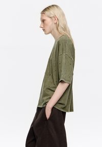 Camiseta oversize verde oliva hecha de tela suave, con tachuelas decorativas negras, mangas cortas y un corte relajado. Combinada con pantalones oscuros.