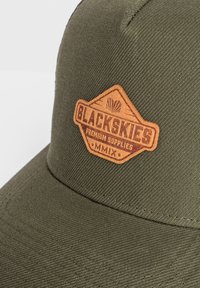 Cappellino da baseball verde con tessuto texturizzato. Presenta una toppa in pelle marrone etichettata "BLACKSKIES", con testo e elementi di design in rilievo.