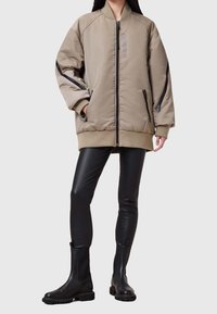 Übergroße beige Bomberjacke mit gerippten Bündchen und Saum, ausgestattet mit schwarzen Reißverschlusstaschen und einem strukturierten Patch auf der Brust. Schwarze Lederleggings und Stiefel.