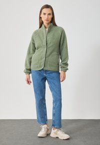 Fleece bunda v jemné světle zelené barvě, s vysokým límcem, předními knoflíky a dvěma bočními kapsami. V kombinaci s modrými džíny a béžovými teniskami.