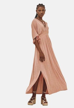 Vestido maxi de suave tela rosa con escote en V, mangas cortas con flecos y una abertura lateral. Lleva sandalias de plataforma.