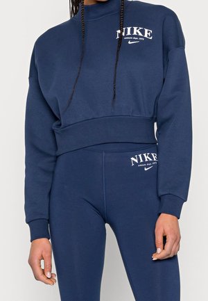 Woman wearing navy blue Nike sweatshirt and leggings with white logo and text "Athletic dept. 1972," showing upper body and hips.