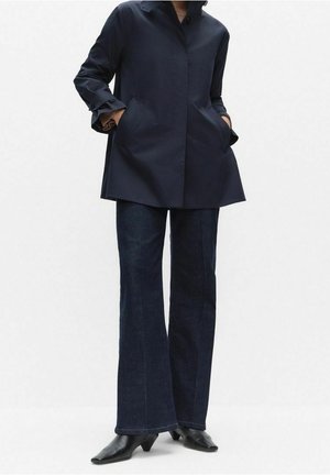 Personne portant un manteau bleu marine avec les mains dans les poches, un jean évasé foncé et des bottines noires à bout pointu, debout devant un fond blanc uni.