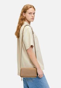Tasche aus hellbraunem Leder mit glatter Oberfläche, minimalistischem Design, Reißverschluss und verstellbarem Schulterriemen, getragen über einem hellen Hemd.