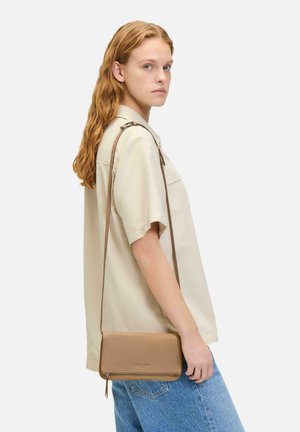 Tasche aus hellbraunem Leder mit glatter Oberfläche, minimalistischem Design, Reißverschluss und verstellbarem Schulterriemen, getragen über einem hellen Hemd.