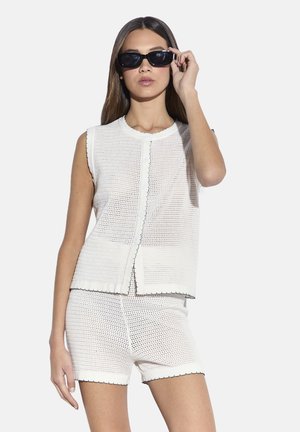 Top senza maniche in maglia bianca e pantaloncini abbinati con una texture traforata. Bordi smerlati e cuciture nere a contrasto. Accessorio occhiali da sole.