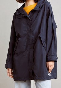 Personne portant une veste de pluie à capuche bleu marine sur un pull moutarde et un jean bleu clair, se tenant devant un fond uni.