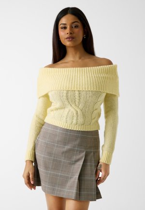 Giovane donna che indossa un maglione a trecce giallo pallido con scollo a barca e una minigonna plissettata marrone a scacchi su sfondo bianco.