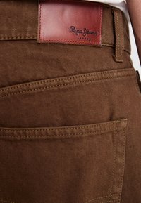 Jeans din denim maroniu cu o finişare texturată, având o etichetă din piele inscripționată "Pepe Jeans London" pe talie și detalii de cusătură.
