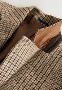 Rakstainā gaiši brūnā un brūnā šaha jaku ar brūnu satīnu oderi, ar ieliektu apkakli un kontrastējošiem auduma elementiem.