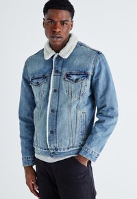 Homme portant une veste en denim bleu clair avec un col en fourrure blanche, une chemise grise en dessous et un pantalon noir, sa main dans sa poche, sur fond blanc.