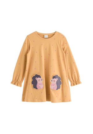 Junges Mädchen mit Brille steht mit gespreizten Beinen, trägt ein gelbes Kleid mit Igel-Aufnähern, blaue Jeans und pinke Sneakers auf einfarbigem Hintergrund.