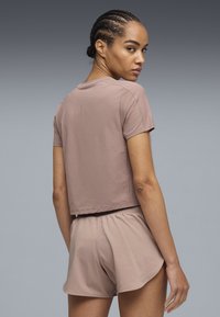 Γυναίκα με πλεξούδα στα μαλλιά, που φοράει ένα ταιριαστό μωβ τοπ με κοντά μανίκια και σορτς, κοιτάζοντας πάνω από τον ώμο της μπροστά σε ένα γκρίζο φόντο με διαβαθμίσεις.