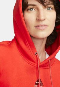 Sudadera roja con cordón, con un logo en el pecho. El material tiene una textura suave y la capucha se ajusta ceñidamente.