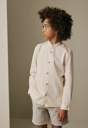 Niño con cabello rizado, vestido con una camisa beige clara abotonada y pantalones cortos a juego, de pie con las manos en los bolsillos, mirando hacia un lado.