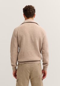 Beige gerippter Strickpullover mit weitem Kragen und langen Ärmeln, kombiniert mit hellbeigen Cargo-Hosen, von hinten gezeigt.