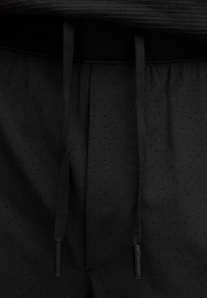 Schwarze Sportshorts mit einer perforierten Oberfläche und flachen Kordeln. Der Bund ist elastisch und sorgt für einen engen Sitz.