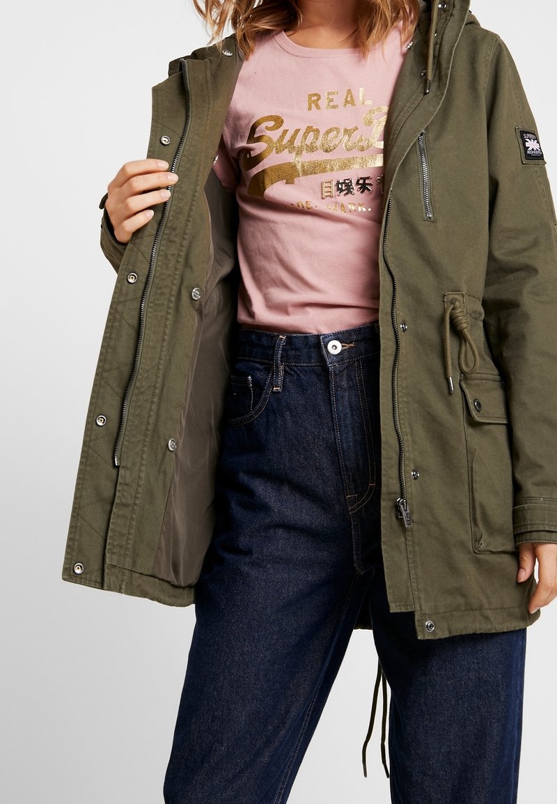 Femme portant une parka vert olive avec fermeture éclair et boutons, un t-shirt graphique rose avec texte doré, et un jean taille haute bleu foncé.