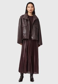 Donna che indossa una giacca di pelle oversized marrone scuro, una gonna plissettata abbinata, un maglione scuro e stivaletti neri su uno sfondo neutro.