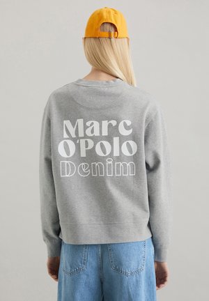 Person med langt blondt hår iført en gul kasket, grå sweatshirt med teksten "Marc O'Polo Denim" og blå cowboybukser set bagfra.