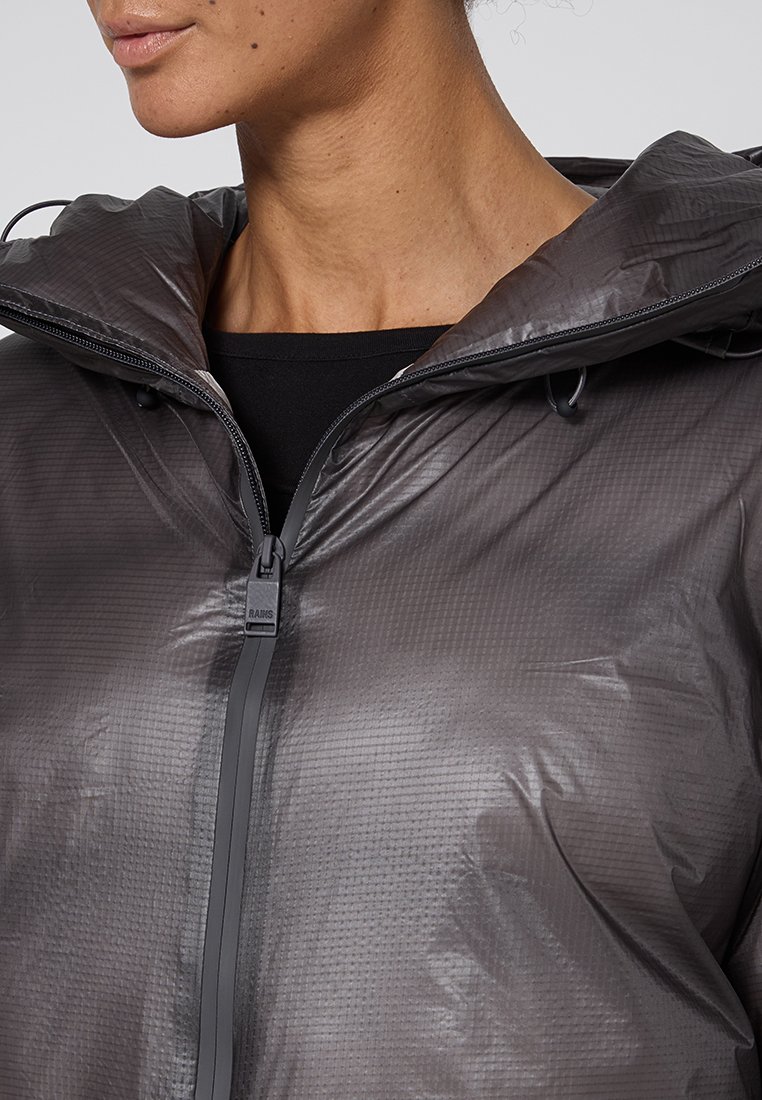 Primo piano di una persona che indossa un'impermeabile grigio scuro e lucido con cappuccio, con la zip anteriore parzialmente chiusa su una maglietta nera.