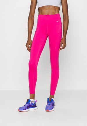 Lyserøde højtaljede leggings med en glat overflade, der har sidelommer og flade syninger. Båret med blå og lilla træningssko.