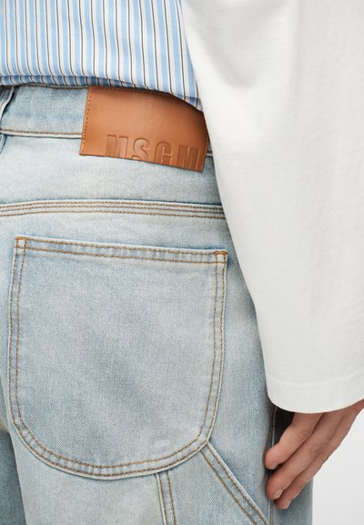 Jean en denim bleu clair avec un patch en cuir marron portant l'étiquette "MSGM", porté avec une chemise à rayures bleues et un haut blanc à manches longues.